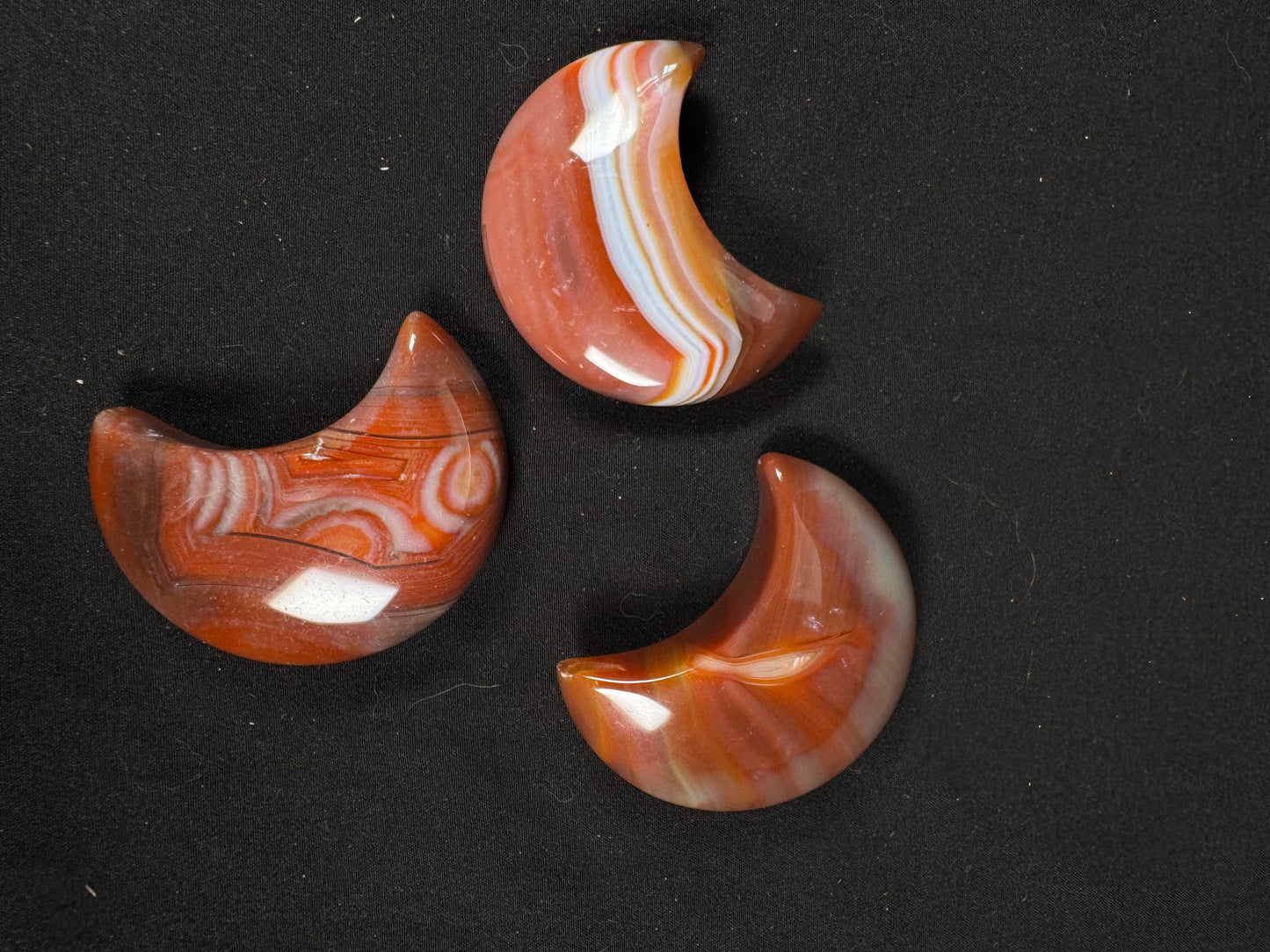 Three polished sardonyx crescent moon carvings displayed on a black fabric background. Each moon features unique natural banding in warm shades of reddish-brown, orange, cream, and white. One moon has bold white and orange stripes, another showcases intricate circular and layered patterns, and the third has soft flowing bands with translucent gray edges. The glossy finish highlights the distinctive colors and patterns of each individual carving.