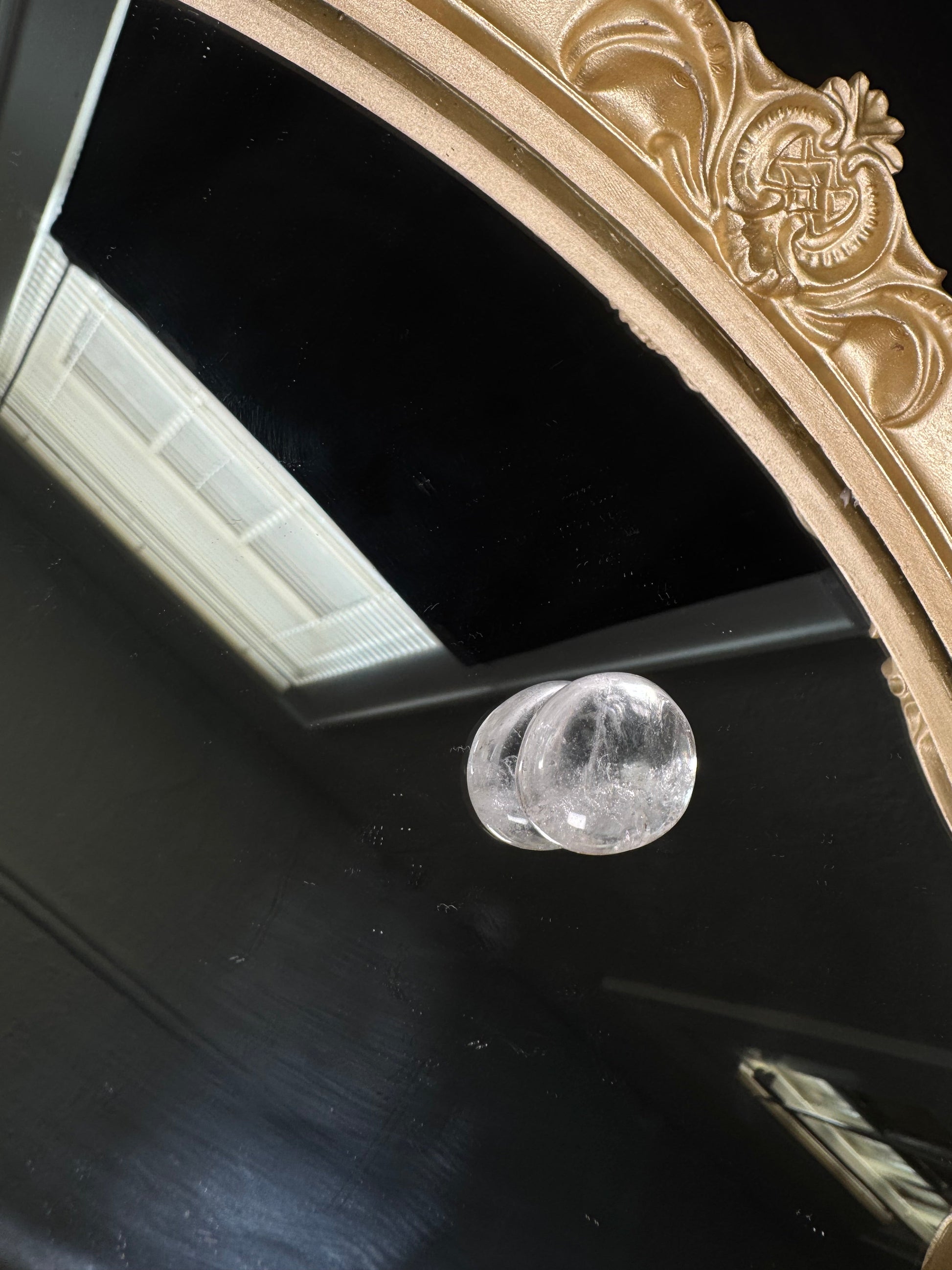 A polished clear quartz tumble rests on a glossy black reflective surface beneath an ornate antique-style gold mirror frame. The crystal is highly transparent with natural internal veils and inclusions that catch the light, showcasing its smooth, rounded shape and glass-like finish.