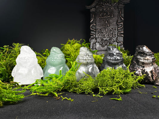 Five crystal Oogie Boogie carvings displayed on vibrant green moss against a black background. The carvings feature a variety of stones, including clear quartz, green aventurine, labradorite, black obsidian, and rhodonite, with a decorative “Rest in Peace” tombstone prop in the background for a spooky, Halloween-inspired display.