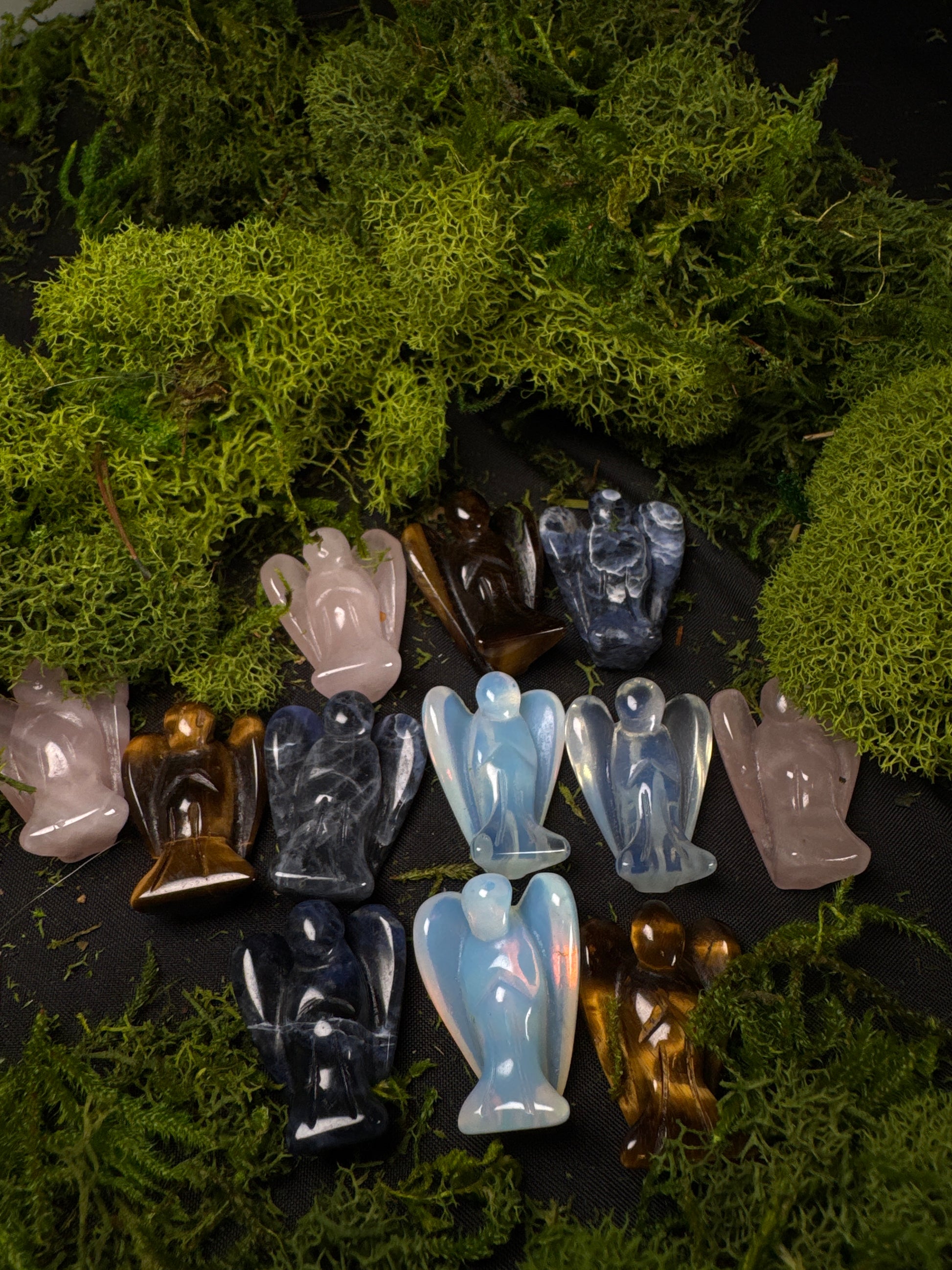 A collection of mini crystal angel carvings displayed on a black fabric backdrop surrounded by lush green preserved moss. The assortment includes a variety of gemstones, such as rose quartz, tiger’s eye, sodalite, blue lace agate, and opalite. Each angel is polished with carved wings and a smooth, glossy finish, showcasing unique natural colors and patterns ranging from soft pinks and blues to rich golden browns and deep navy hues. The moss creates a woodland-inspired display that highlights the beauty of e