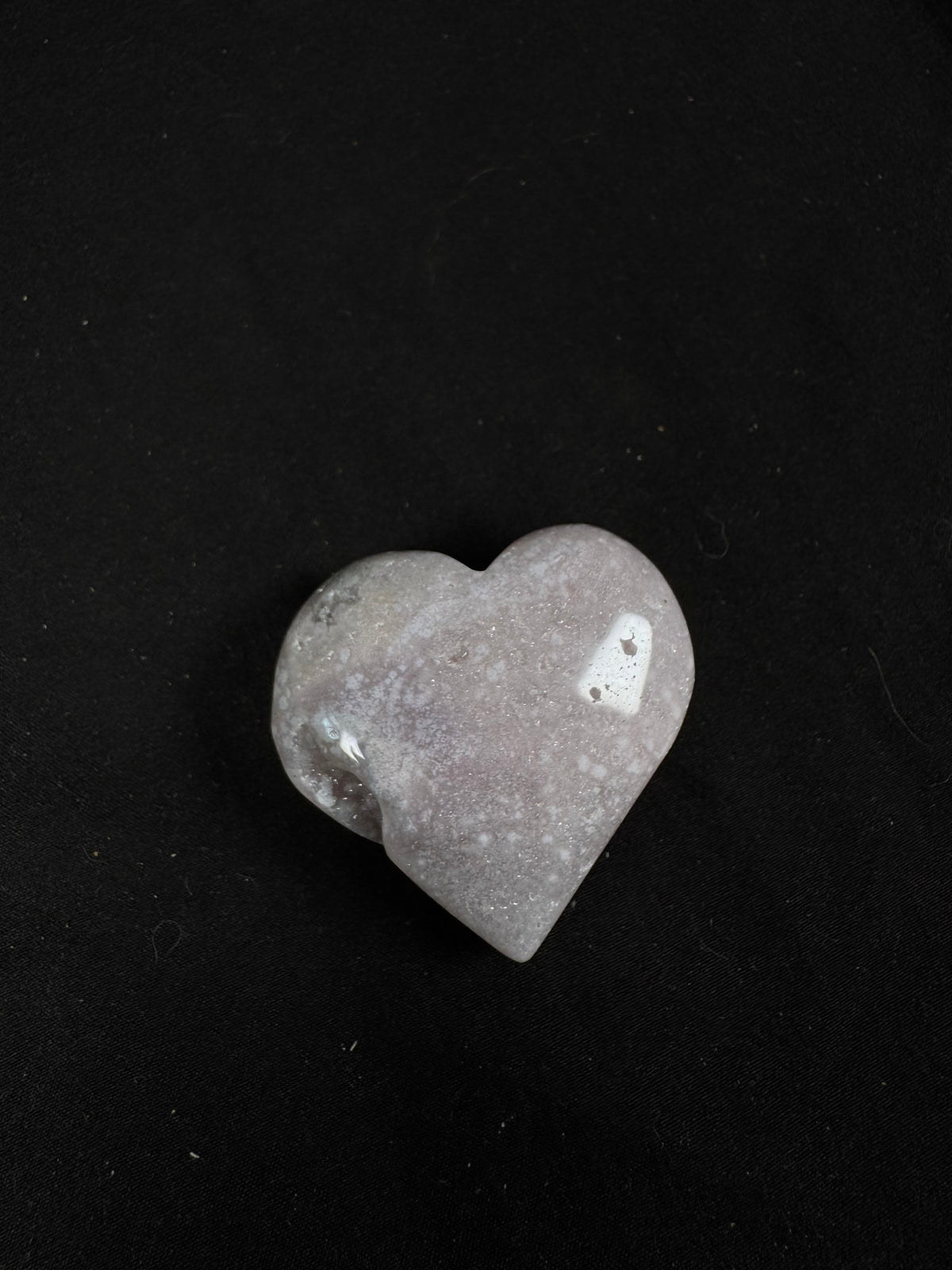 Polished pink amethyst heart featuring soft blush pink, cream, and pale gray tones with subtle druzy sparkle across the surface. The heart has a smooth, glossy finish with a small natural druzy pocket along one side, photographed against a black background.