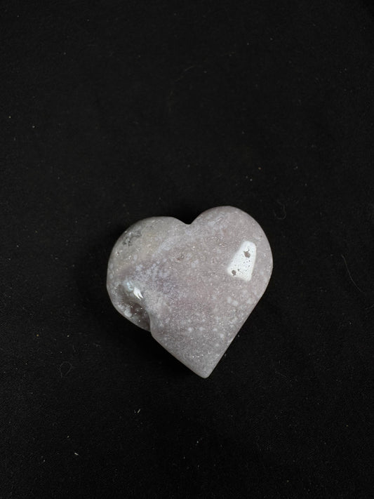 Polished pink amethyst heart featuring soft blush pink, cream, and pale gray tones with subtle druzy sparkle across the surface. The heart has a smooth, glossy finish with a small natural druzy pocket along one side, photographed against a black background.