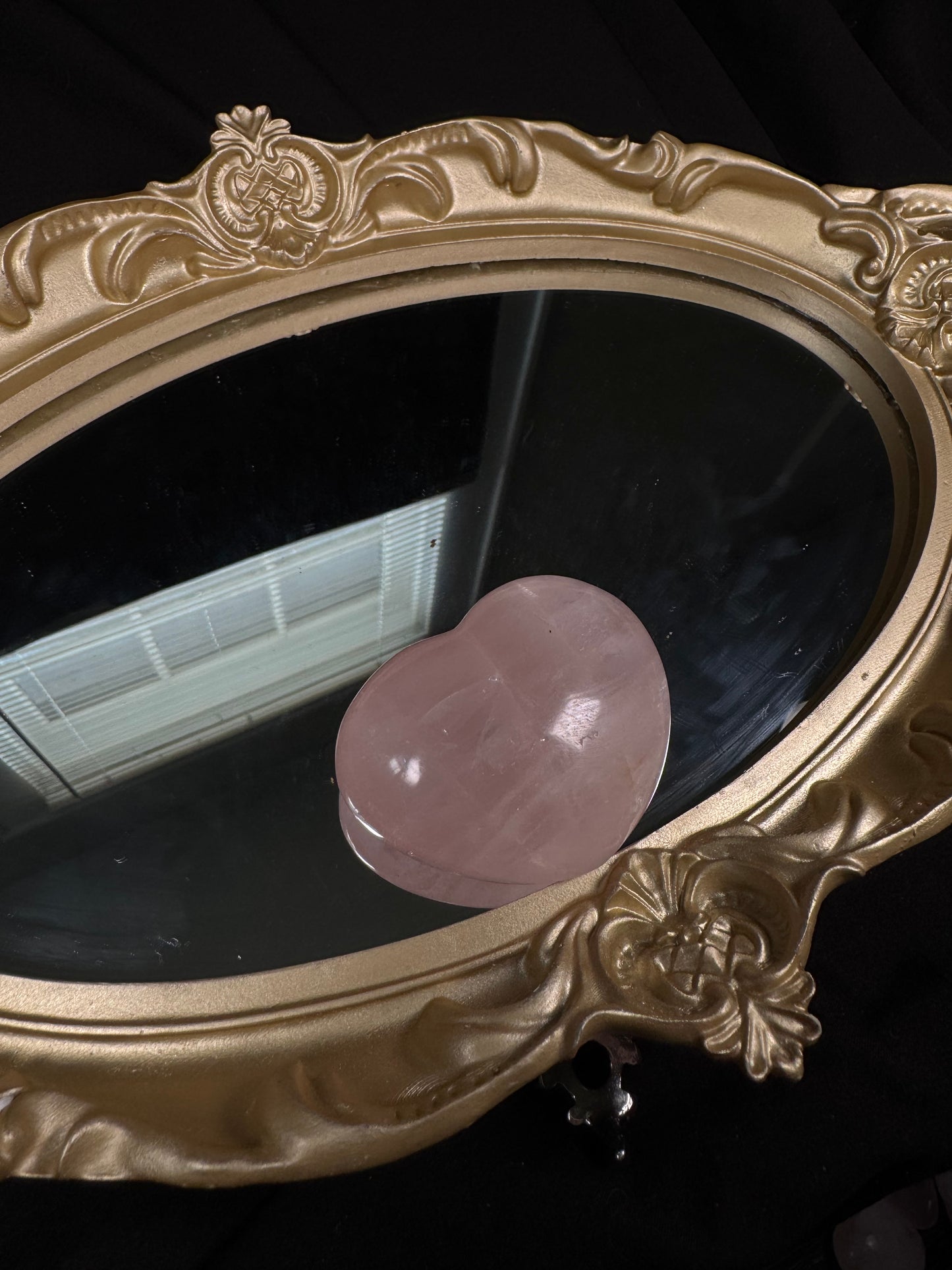 A single polished rose quartz heart resting on an antique-style gold mirror. The heart features a delicate blush pink color with translucent areas, natural veining, and a smooth, reflective surface highlighted by soft lighting.